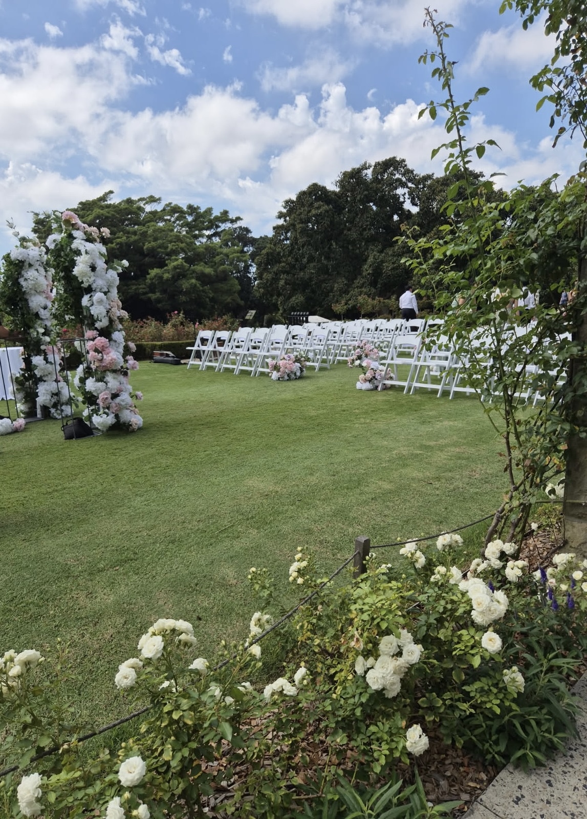Outdoor wedding ceremony setup at the Royal Botanic Garden Sydney with white folding chairs, floral arbour and garden views by Budget Event Hire